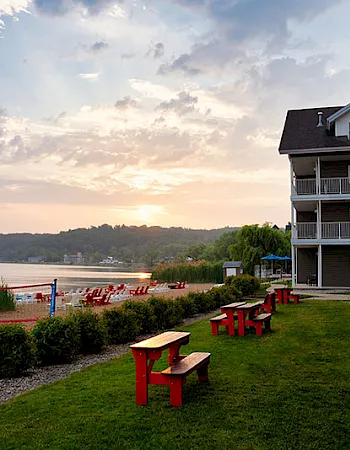 Lakeside resort buildings with manicured grass, red picnic tables, and a dock by the water under a cloudy sunset.