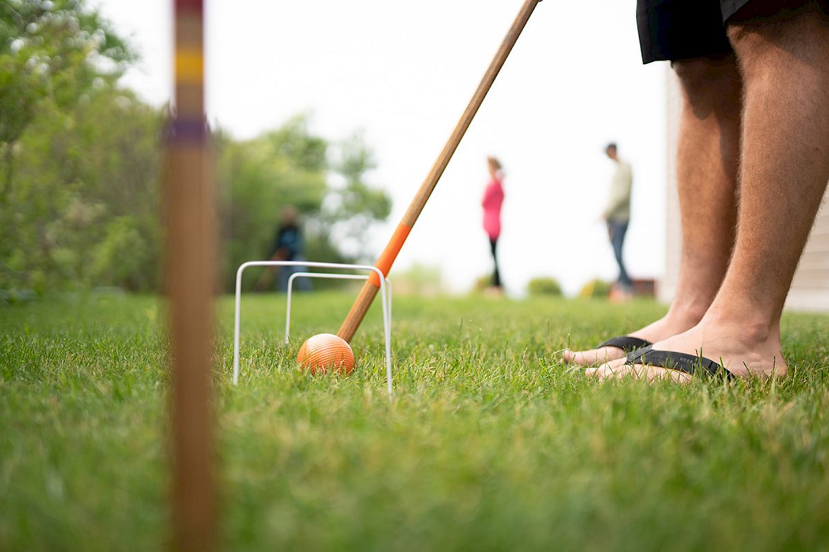 A lawn cricket scene: an orange ball and bat on the grass with players blurred in the background.