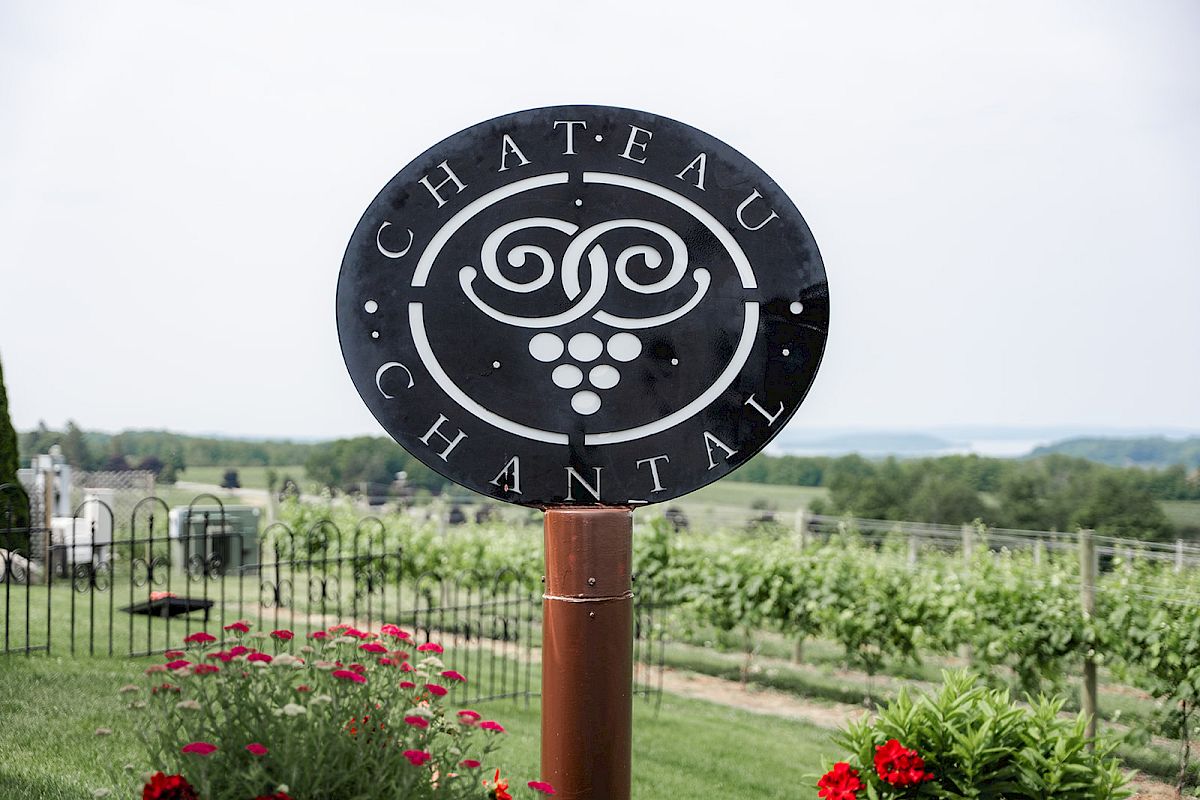 A circular metal sign reading “CHATEAU HANTAL” beside a vineyard garden with flowers and a fence.