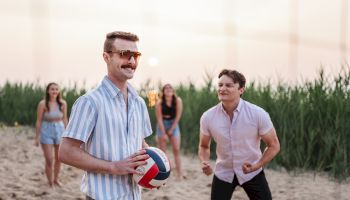 Two men playing beach volleyball with a ball; others watch in the background on a sunny beach. The man foreground wears striped shirt, sunglasses.