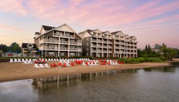 A beachfront resort with multi-story buildings, numerous red and white lounge chairs on the sandy shore, and calm water at sunset.