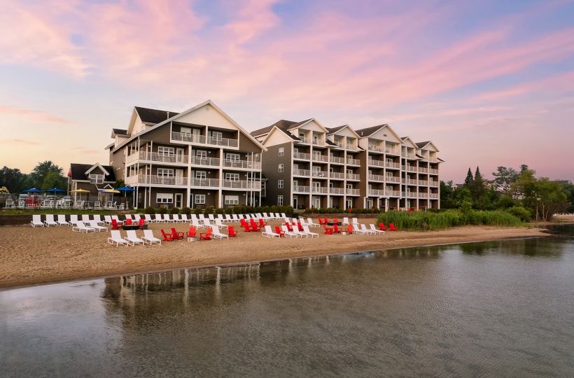 A beachfront resort with multi-story buildings, numerous red and white lounge chairs on the sandy shore, and calm water at sunset.