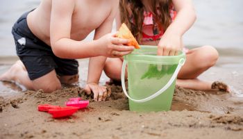 Two children play on the beach, using a green bucket and red toy boats in the sand by the water.