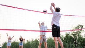 People playing beach volleyball near greenery, one person jumping to spike the ball while others watch or prepare to play.