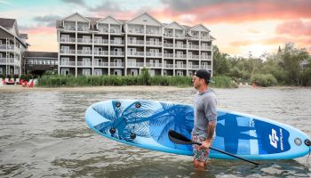 A man stands in water holding a blue paddleboard near a multi-story building at sunset, with greenery and red chairs in the background.
