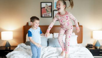 Two children joyfully jump on a bed, holding hands, in a cozy room with lamps and pictures on the wall.
