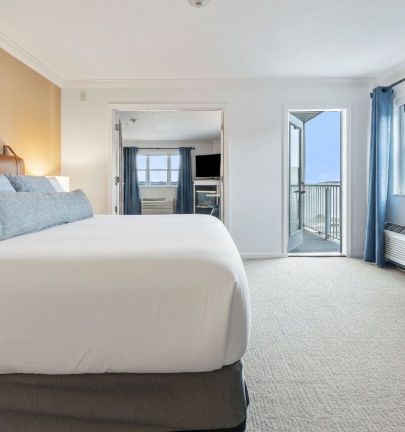 A bright hotel-style bedroom with a large white bed, beige walls, blue curtains, a dresser, and a balcony door opening to a view, all in neat, modern decor.