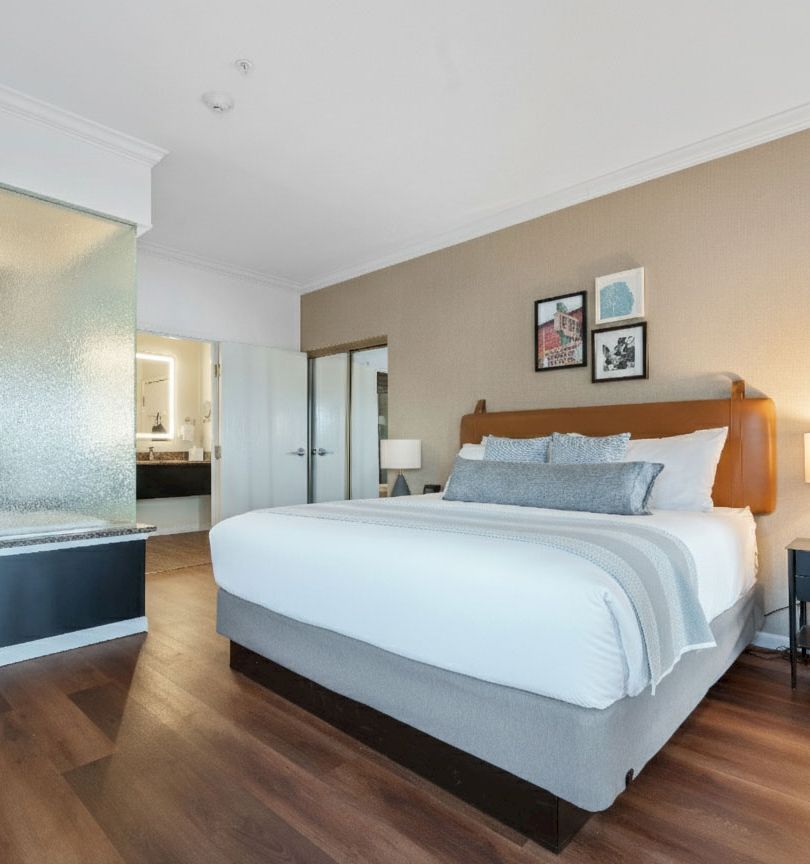 A modern hotel-style bedroom with a king bed, wooden floor, bedside tables, and a frosted glass partition near a bathroom, bathed in warm light.