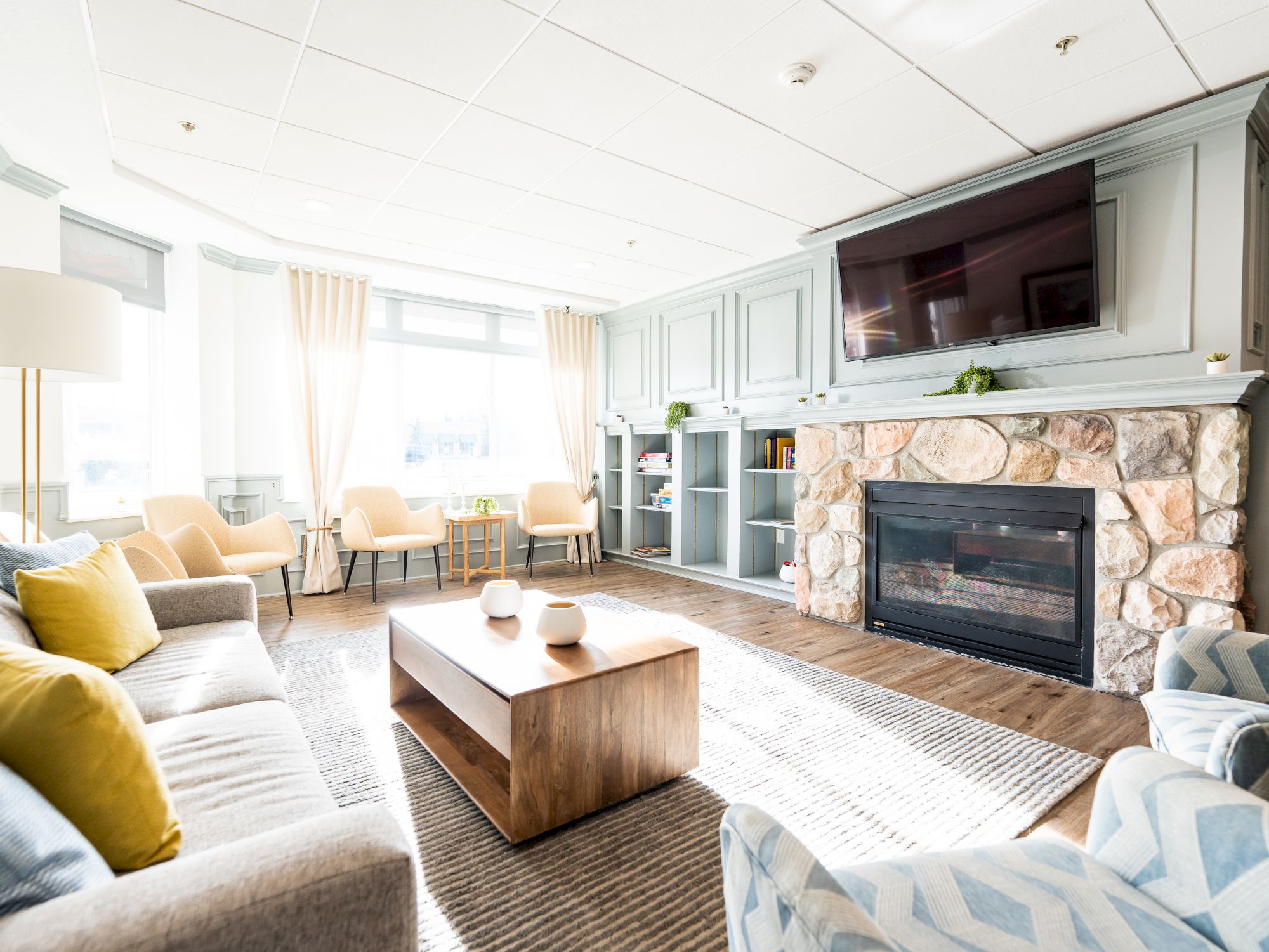 A cozy living room with a fireplace, large TV, sofas, armchairs, coffee table, and bright natural light from a large window, ending the sentence.