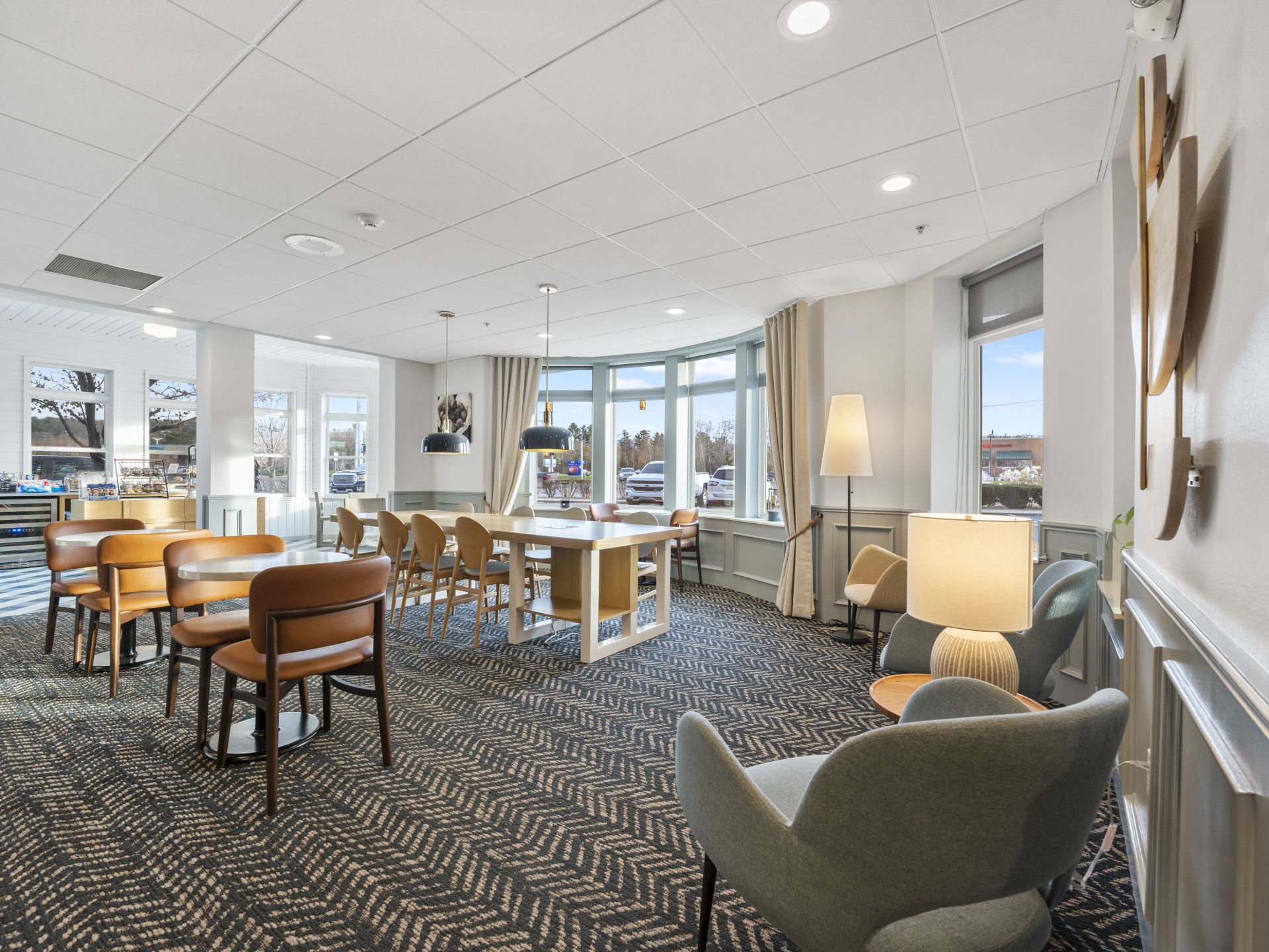 A spacious, well-lit room features wooden tables and chairs, comfortable seating, and large windows allowing natural light.