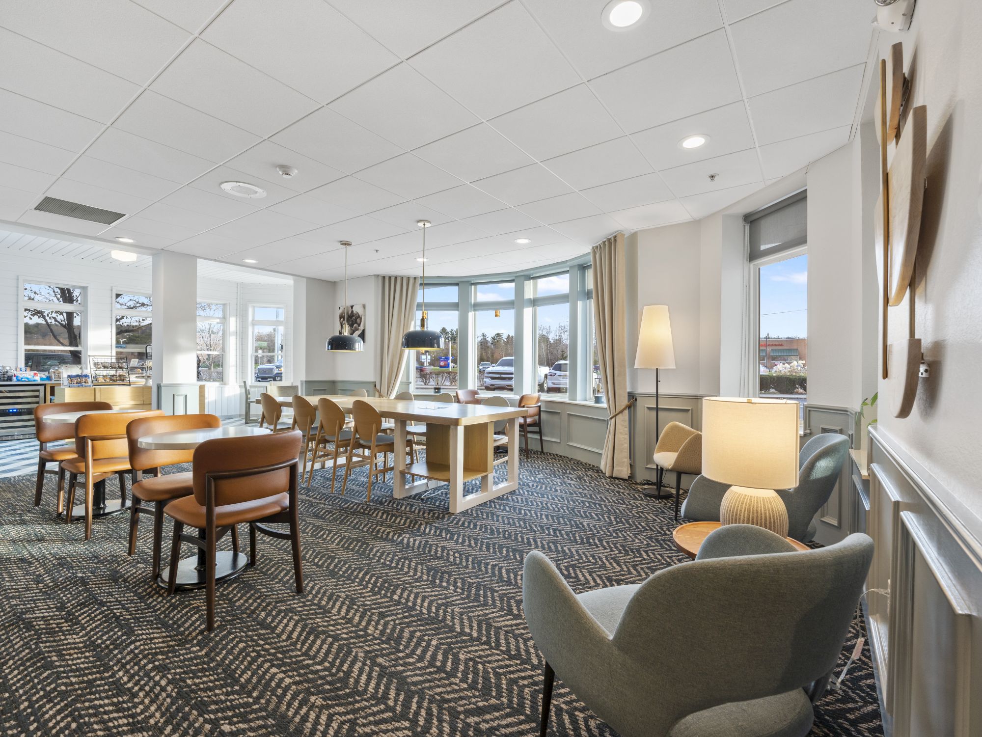 A spacious, well-lit room features wooden tables and chairs, comfortable seating, and large windows allowing natural light.