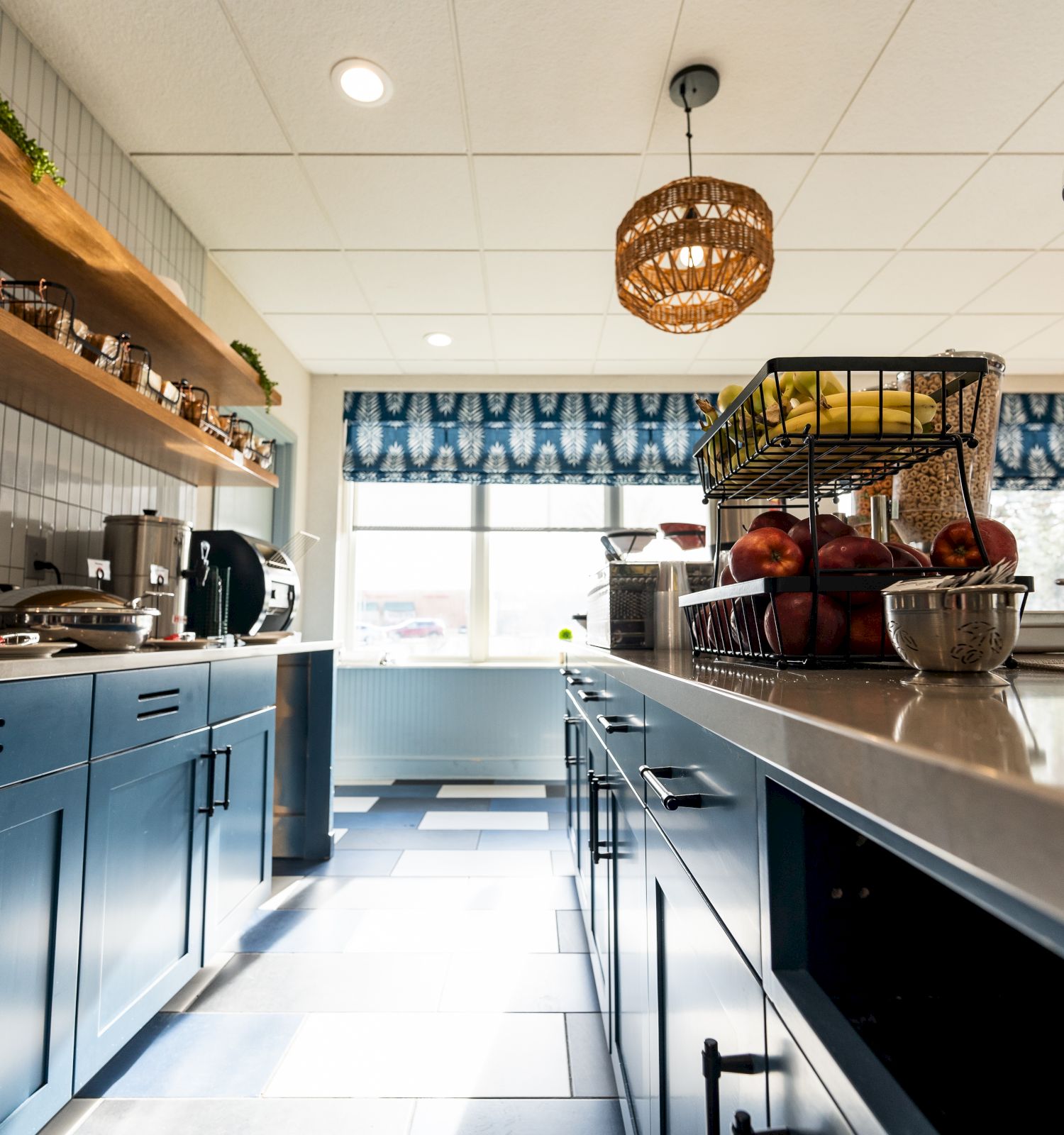 A modern kitchen with blue cabinets, open wooden shelves, various appliances, a basket with fruits, and a woven light fixture hangs from the ceiling.