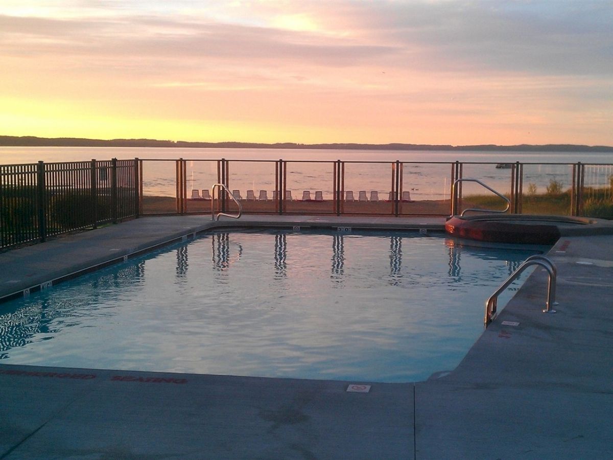 An outdoor pool and hot tub with a fence overlooking a lake at sunset, with chairs arranged along the shoreline in the background.