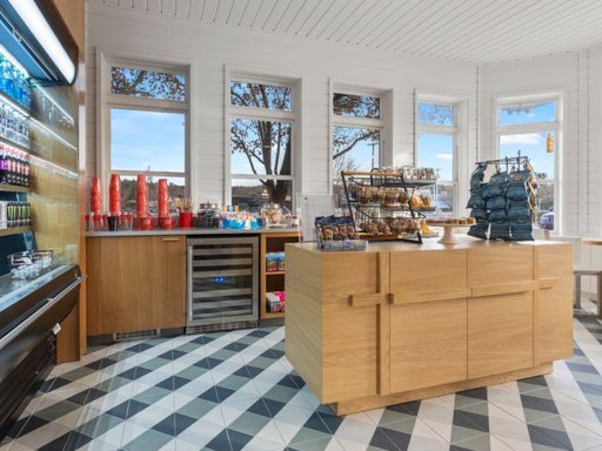 The image shows a bright, modern store with shelves of products, a central wooden counter, geometric tiled floor, and large windows letting in natural light.