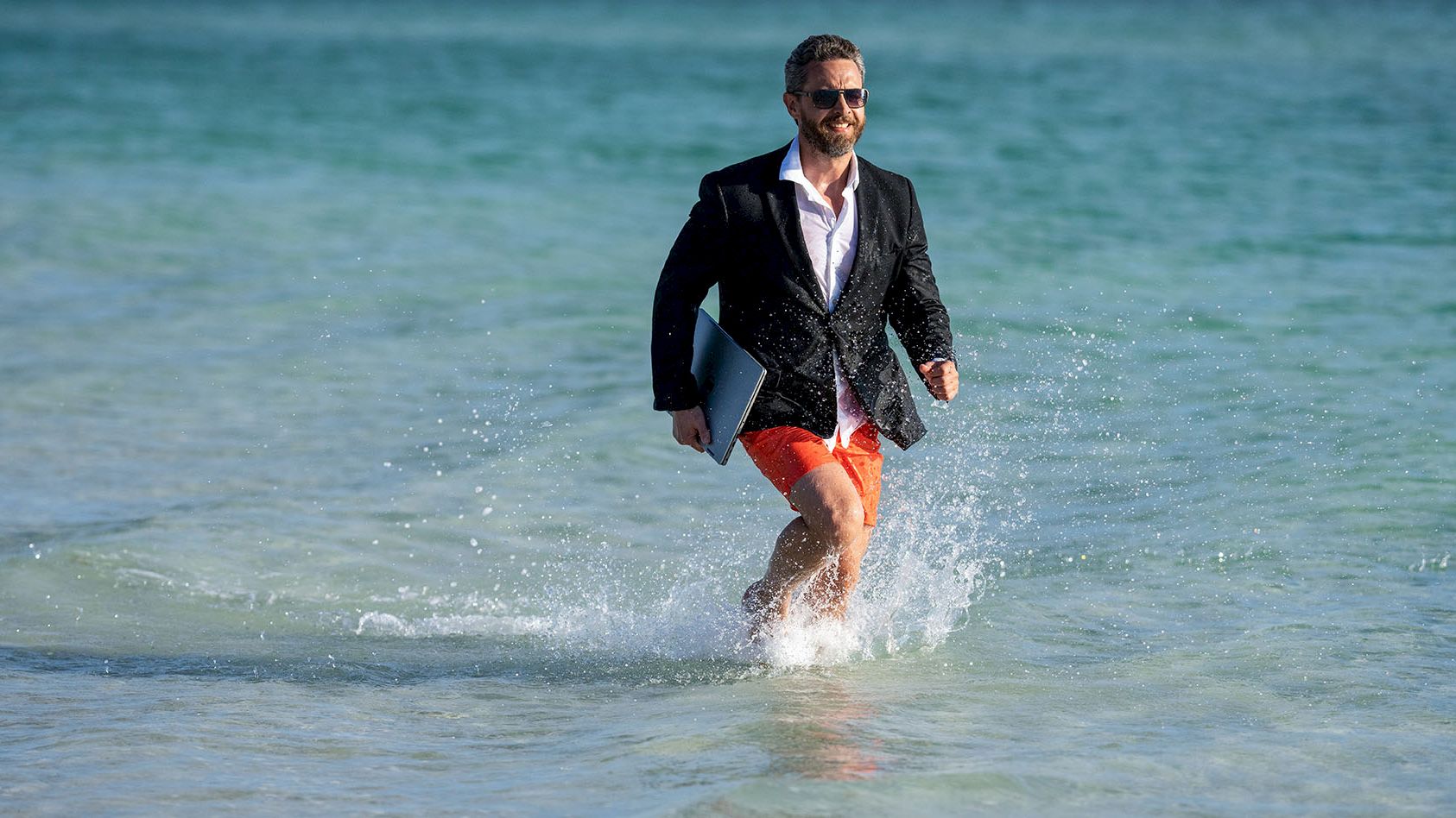 A man in a suit runs along a choppy beach with a surfboard, throwing spray as he smiles at the camera.