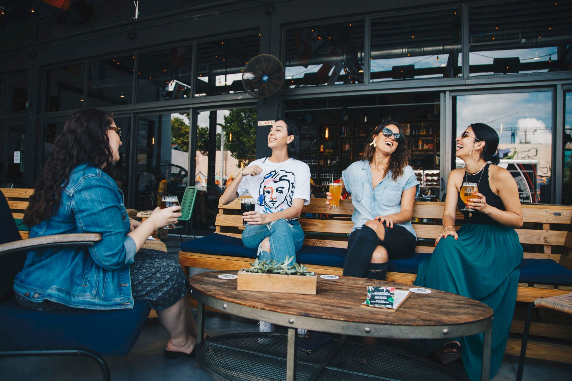 Four people are sitting outdoors at a wooden table, enjoying drinks and laughing together, suggesting a lively social atmosphere.