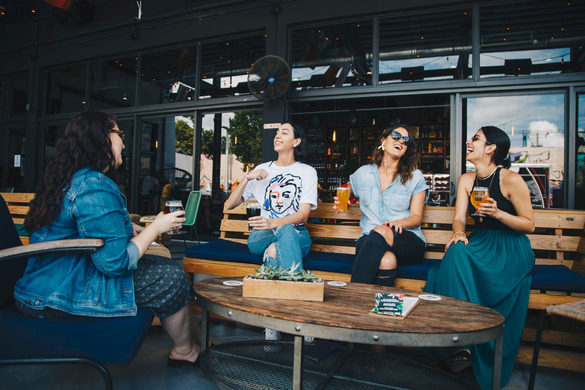 Four people are sitting outdoors at a wooden table, enjoying drinks and laughing together, suggesting a lively social atmosphere.