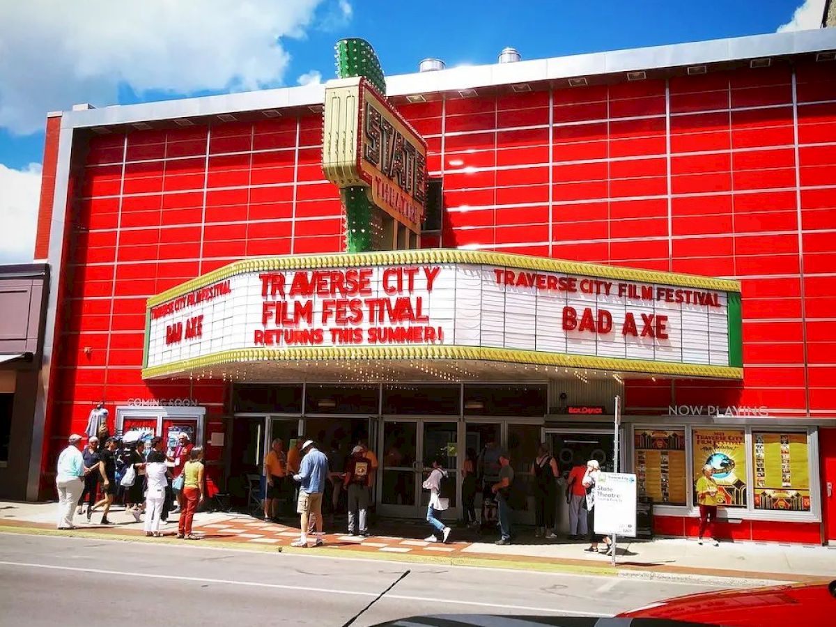 A red theater marquee reads "Traverse City Film Festival, Bad Axe". There are people gathered in front of the theater with a sunny sky above.