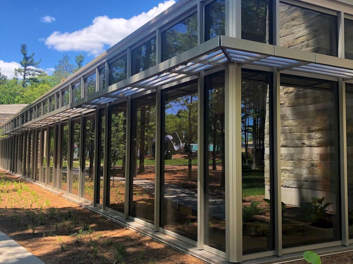 The image shows a modern building with large glass windows and metal framing, set against a clear blue sky with some greenery around it.