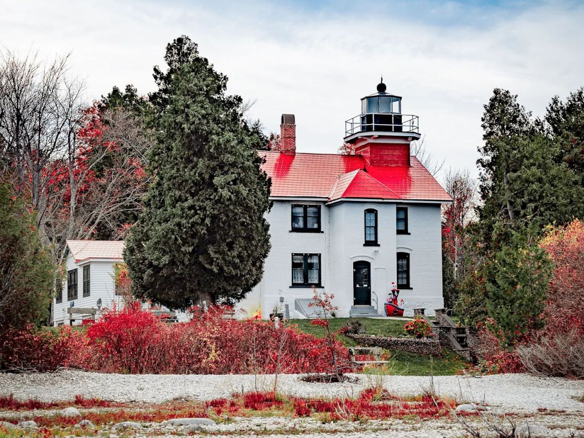 A picturesque lighthouse with a red roof amidst greenery and autumn foliage sits by a rocky shoreline under a partly cloudy sky.