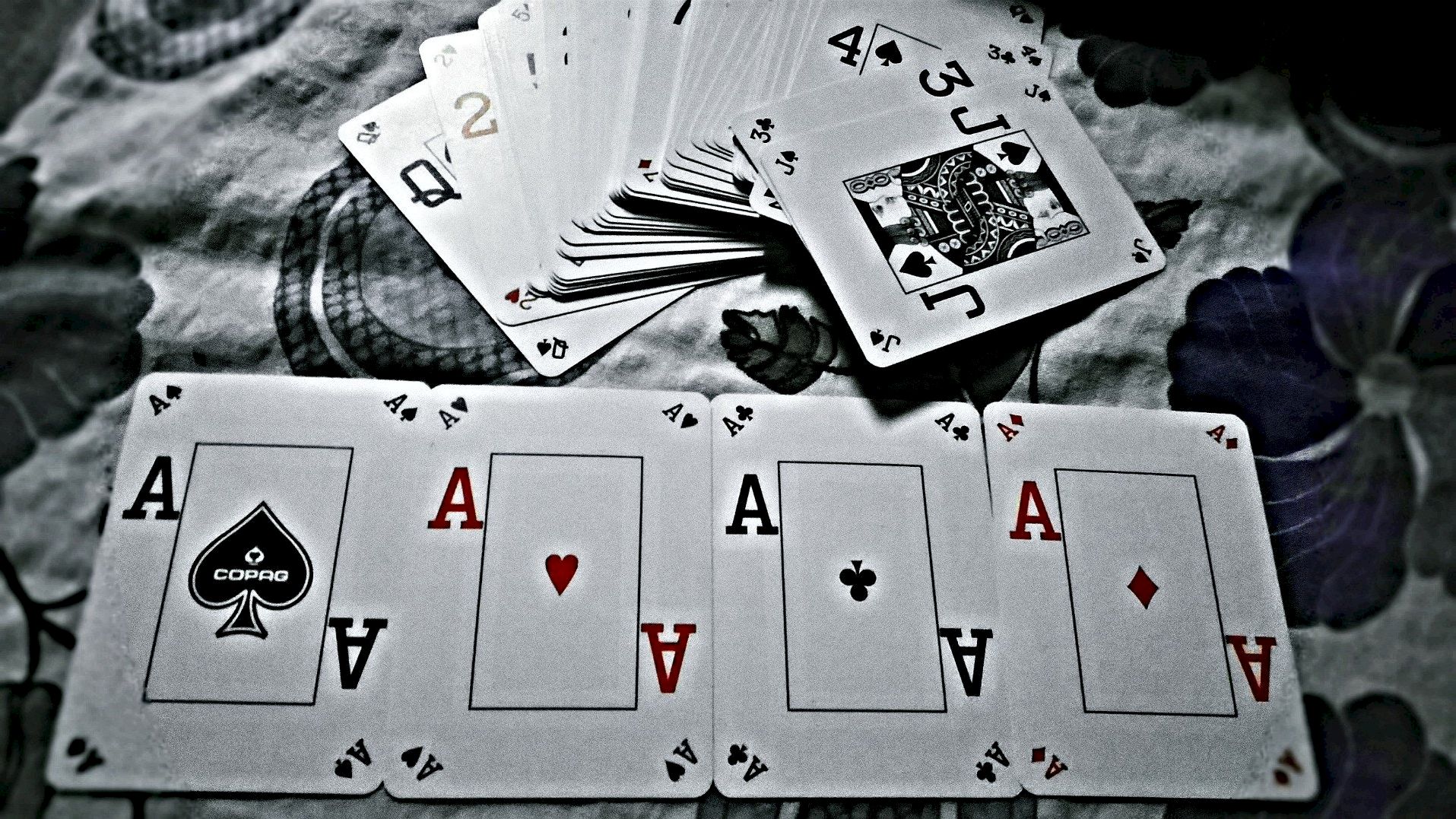 A spread of playing cards is shown, featuring multiple aces in the foreground and a scattered deck in the background, on a floral surface.