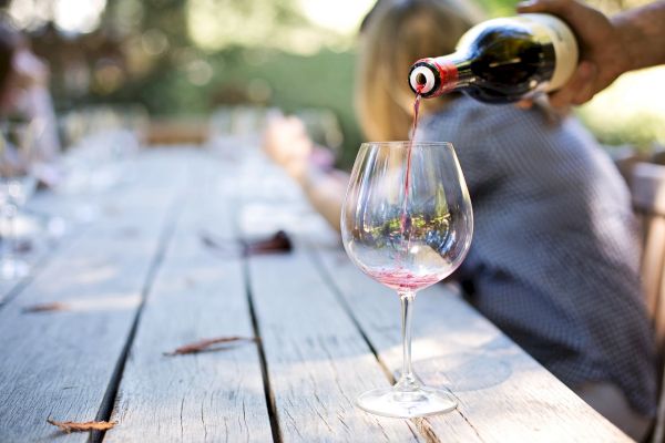 A hand pours red wine into a glass on a wooden outdoor table; the background features people in a relaxed, blurred setting.
