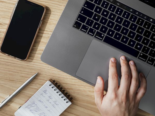 A hand on a laptop trackpad, a smartphone, a pen, and a small notepad with handwritten notes are on a wooden surface.