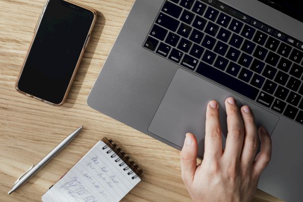 A hand on a laptop trackpad, a smartphone, a pen, and a small notepad with handwritten notes are on a wooden surface.