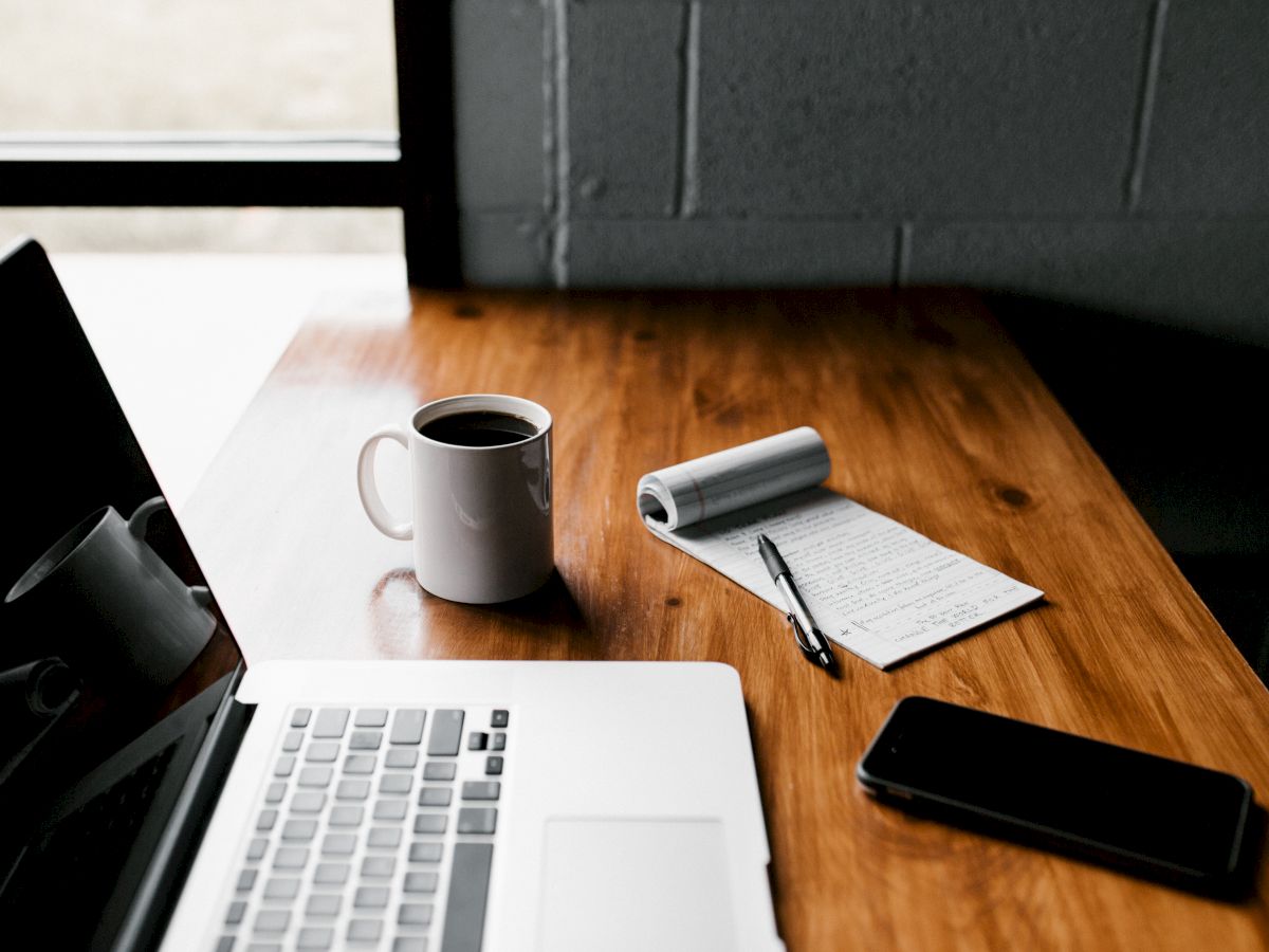 A wooden desk with a laptop, white coffee mug, notebook, pen, and smartphone near a window.