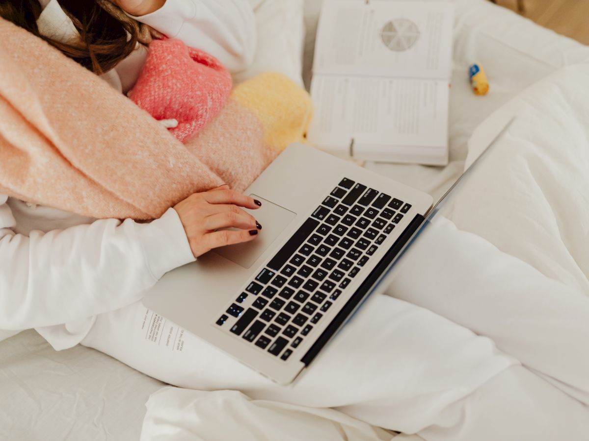 A person with a scarf uses a laptop on a bed, with open books nearby, in a cozy setting.