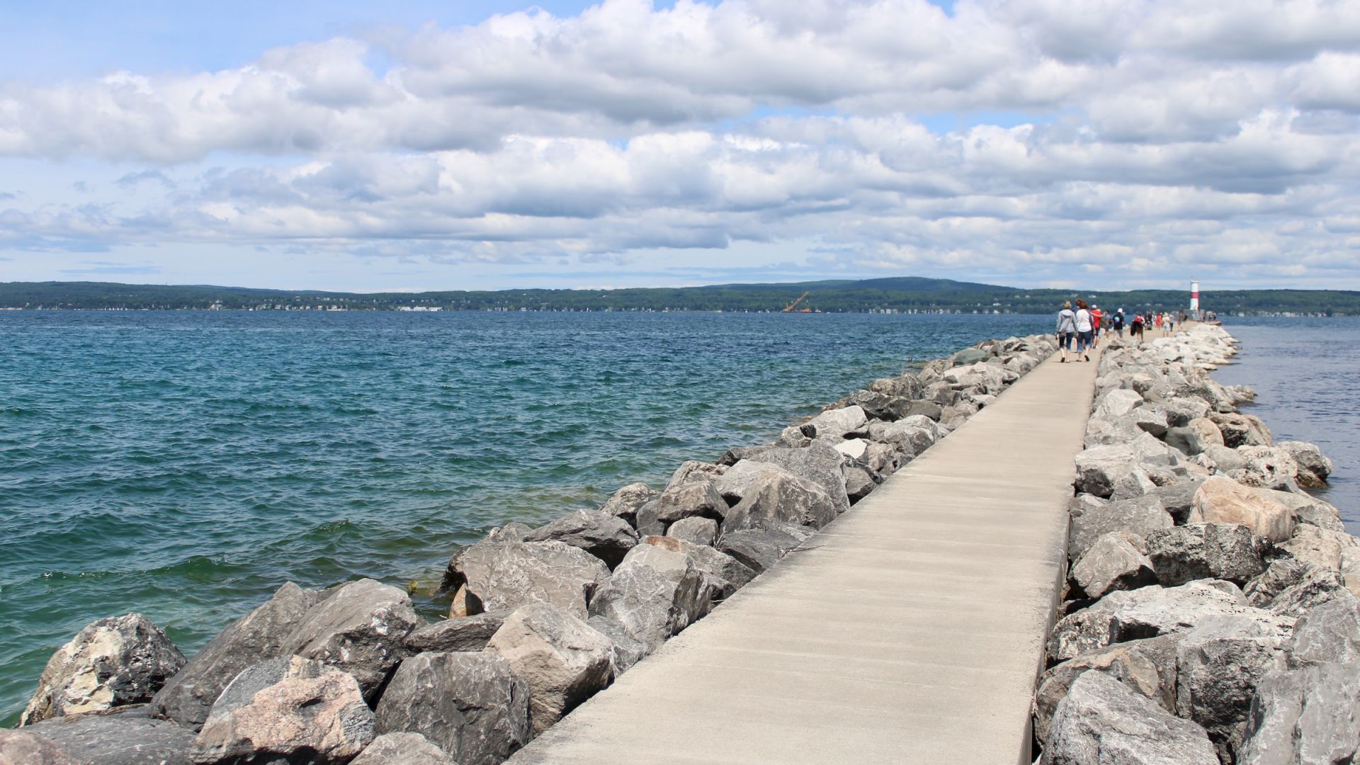 A concrete walkway extends into a body of water, with rocks on either side and people in the distance, under a partly cloudy sky.
