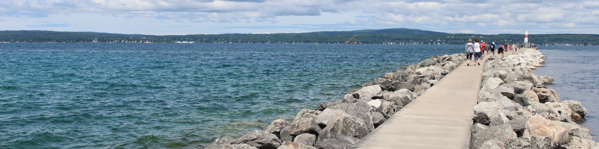 A concrete walkway extends into a body of water, with rocks on either side and people in the distance, under a partly cloudy sky.