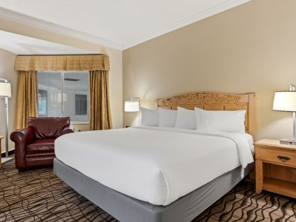 A hotel room with a king-sized bed, white linens, two nightstands with lamps, a brown armchair, a window with curtains, and a small side table.