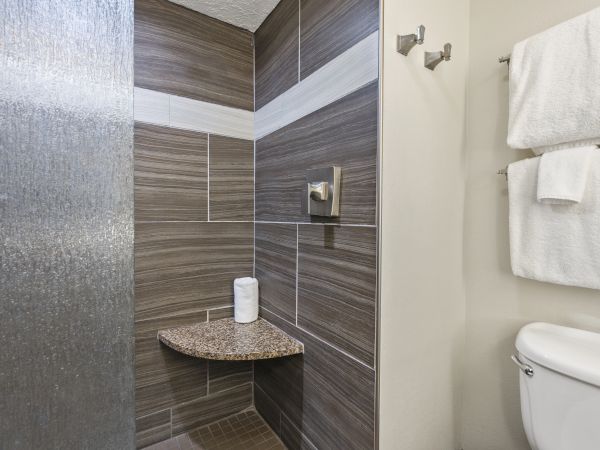 The image shows a modern shower area with grey tiles, a toilet, and towels hanging on the wall. There is also a small granite shelf with tissue paper.