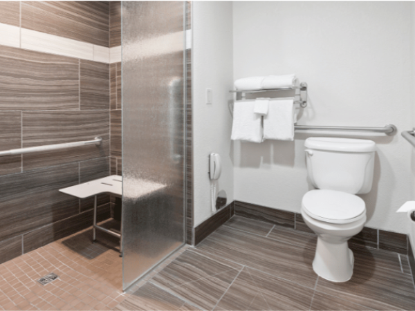 An accessible bathroom with a walk-in shower, grab bars, a wall-mounted foldable shower seat, a toilet, and a towel rack with white towels.