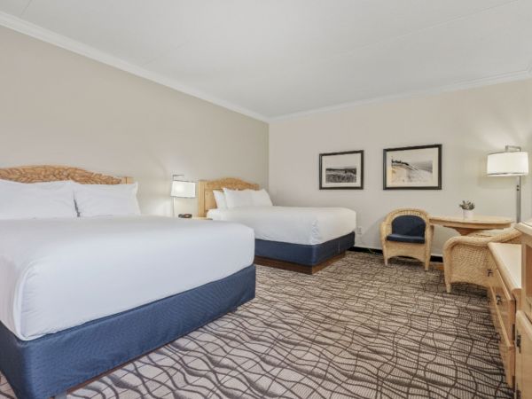 A hotel room with two double beds, a desk with a chair, an additional chair, two framed pictures on the wall, and lamps providing lighting.