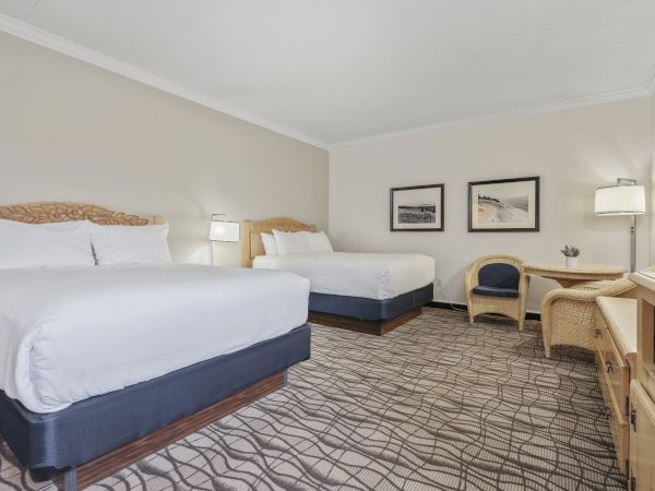 A hotel room with two double beds, a desk with a chair, an additional chair, two framed pictures on the wall, and lamps providing lighting.