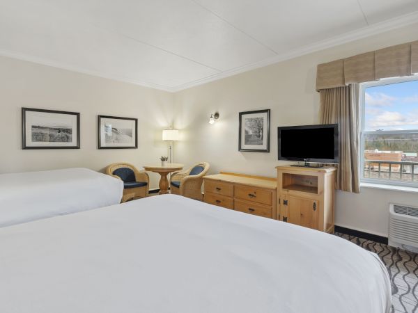 A hotel room with two beds, a TV, dresser, chairs, table, lamps, and a window offering a view outside.