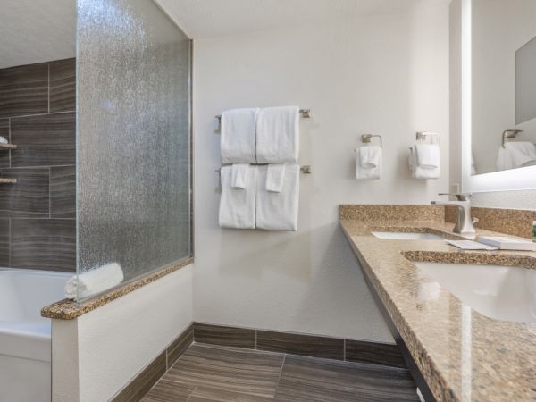 A clean, modern bathroom with a granite countertop, sink, bathtub with glass partition, wall-mounted towel racks with towels, and a mirror.