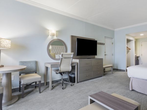 The image shows a modern hotel room with a bed, desk, chair, flat-screen TV, round table with chairs, and minimalistic furnishings, in neutral tones.