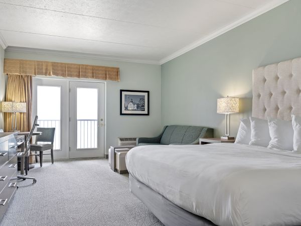 The image shows a modern hotel room with a large bed, a sofa, a desk, a flat-screen TV, and French doors leading to a balcony.