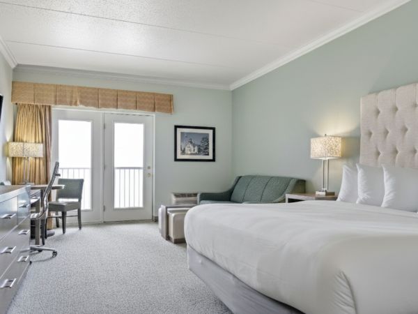 A bright, modern hotel room featuring a king-sized bed, a cozy seating area, and doors leading to a balcony.