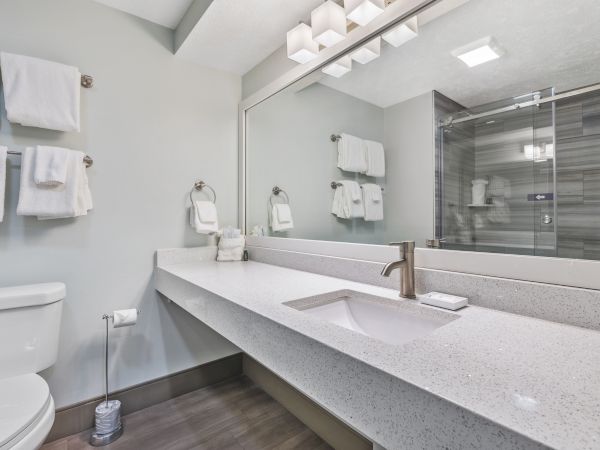 A modern bathroom with a large mirror, white countertop, sink, toilet, and towels on racks. A glass-enclosed shower is visible in the background.