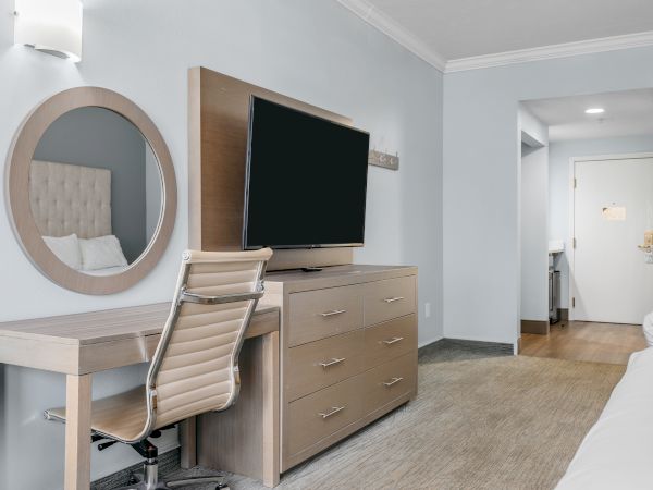 The image features a modern hotel room with a desk, a chair, a large mirror, a flat-screen TV on a dresser, and two beds.