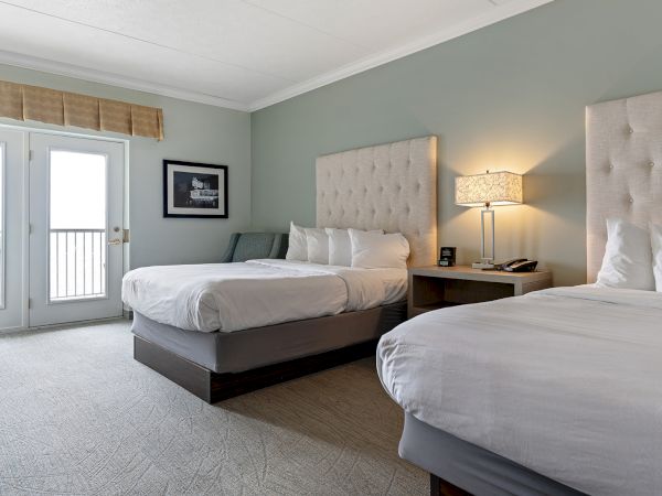 The image shows a modern hotel room with two double beds, a nightstand with a lamp, a chair, and doors leading to a balcony.