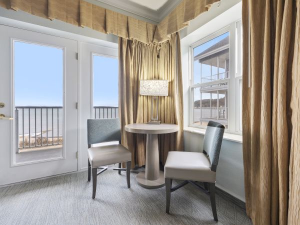 A small cozy room with two chairs and a round table by the window, featuring beige curtains and access to an outdoor view through a glass door.