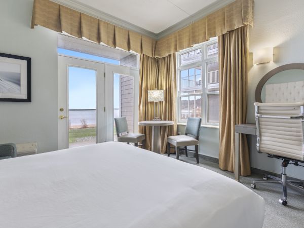 A bright, modern hotel room with a bed, desk, and chairs. It has a large window and doors opening to an outdoor view.