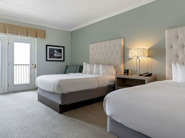 A hotel room with two beds, a nightstand with a lamp, a patio door, an armchair, and a picture on the wall above the chair.