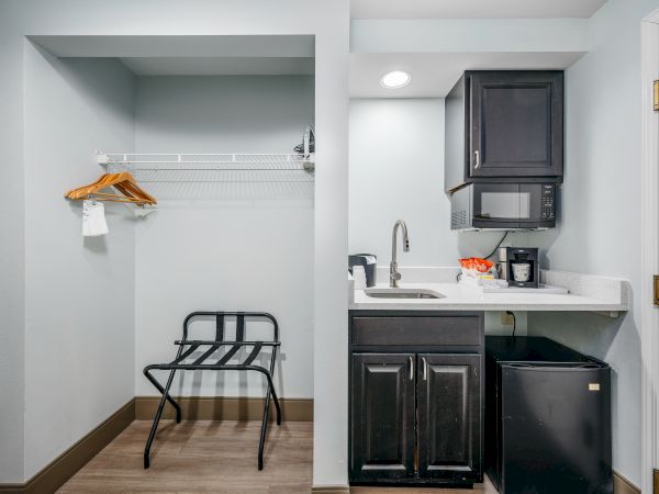 The image shows a small hotel kitchenette area with a sink, microwave, mini-fridge, a coffee maker, and a closet space with hangers and a luggage rack.