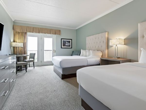 A hotel room with two beds, a TV, a dresser, a desk, a lamp on a nightstand, and a balcony with a view.